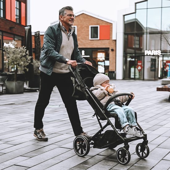 Man met bril en groene jas duwt een zwarte Deryan buggy met een baby erin, buiten.