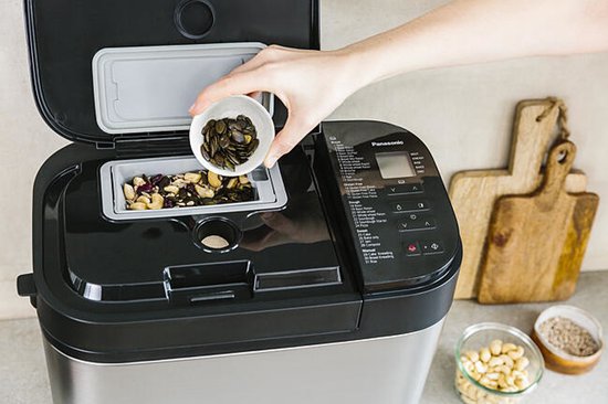 Zilverkleurige broodbakmachine met deurtje open, een hand voegt ingrediënten toe aan de broodvorm.