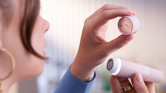 Vrouw houdt een roze Philips gezichtshaarverwijderaar en een kleine spiegel vast, om zichzelf te bekijken.