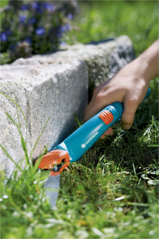Hand maait gras langs stenen muur met blauwe Gardena graschaar.