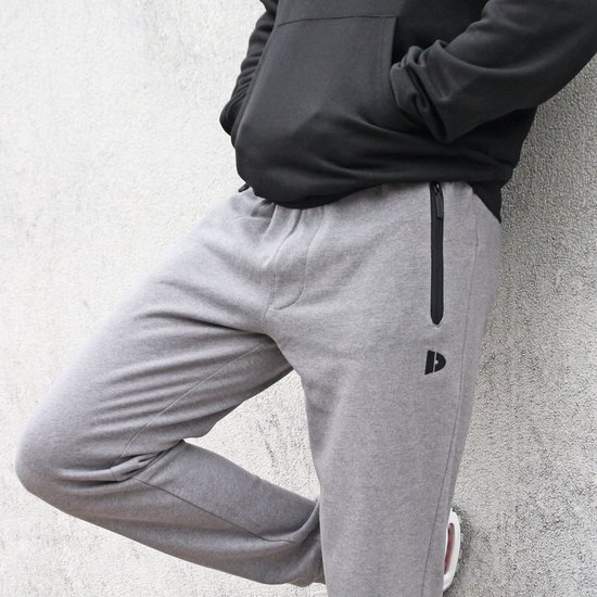Man leunt tegen een muur in een grijze joggingbroek met ritssluiting op de heup.
