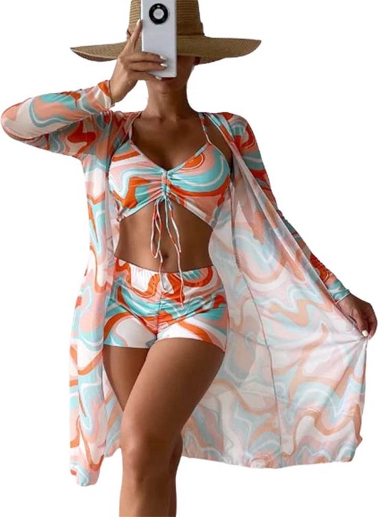 Vrouw staat met een telefoon in de hand, draagt een 3-delige strandset met oranje, blauwe en witte golvende print. Met bikini top, short en kimono.