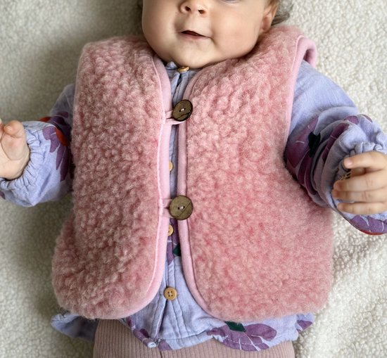 Baby met roze wollen gilet, paars bloemenprint shirtje en roze broekje.
