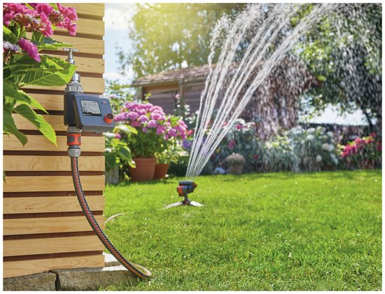 Gardena besproeiingscomputer, aangesloten op een kraan, met sproeiende sprinkler in de tuin.