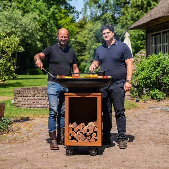 Twee mannen bij een grote zwarte grill op cortenstaal onderstel met houtopslag, in een tuin.