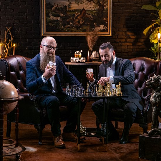 Twee mannen met baarden spelen schaak. Een houdt whiskyglas met wereldbol opdruk.