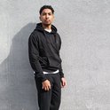 Man staat voor een muur, draagt een zwarte hoodie en een zwarte joggingbroek.