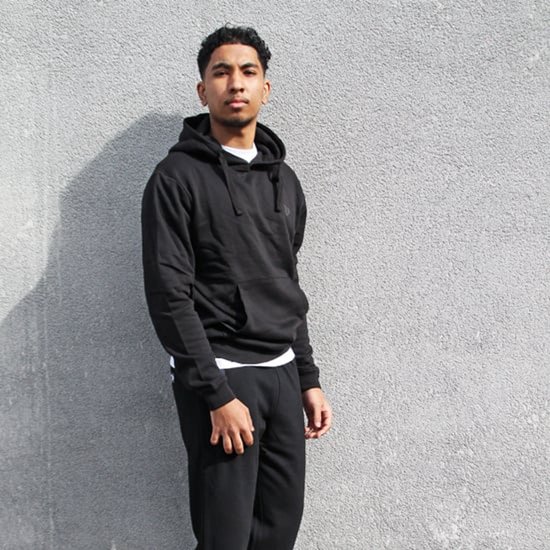 Man staat voor een muur, draagt een zwarte hoodie en een zwarte joggingbroek.