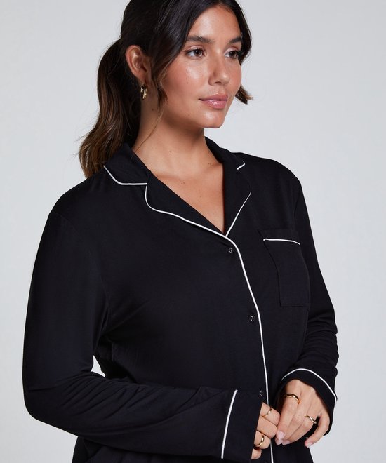 Model met donker haar in een staart draagt een zwart pyjamashirt met witte biezen en opgestikte borstzak.