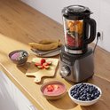 Blender op houten aanrecht met glazen kan vol fruit, aardbeien, blauwe bessen en twee smoothie bowls.