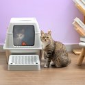 Grijze top entry kattenbak met een grijze tabby kat ervoor. Een tweede kat kijkt uit het raam van de bak. In een woonkamer setting.