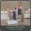 Een jong meisje met grijze jurk en roze shirt hangt poppenkleding aan een wit kleerhangerrek. Voor kinderen vanaf 3 jaar.