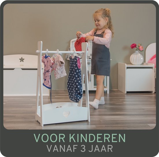 Een jong meisje met grijze jurk en roze shirt hangt poppenkleding aan een wit kleerhangerrek. Voor kinderen vanaf 3 jaar.