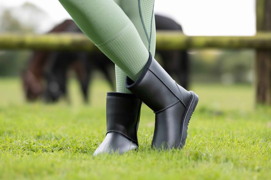Bottes noires avec legging vert sur l'herbe, avec un cheval en arrière-plan.