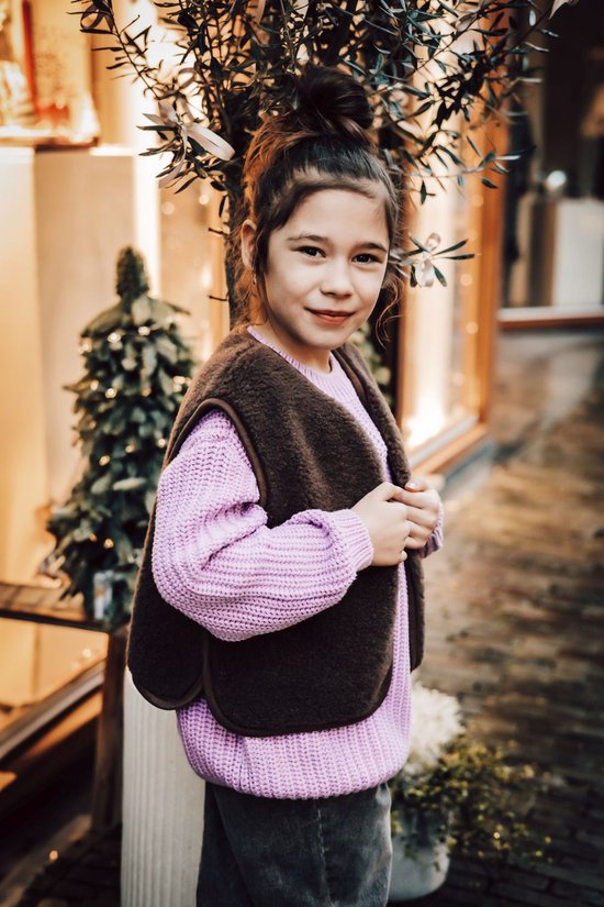 Meisje met opgestoken haar, glimlachend, draagt een bruin teddy fleece gilet en paarse trui.