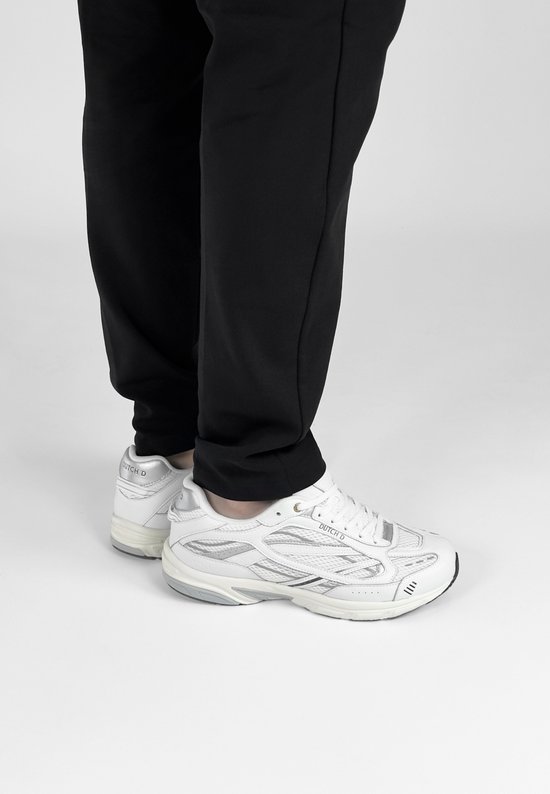 Persoon draagt witte "DUTCH'D" sneakers met zilveren accenten en zwarte broek.