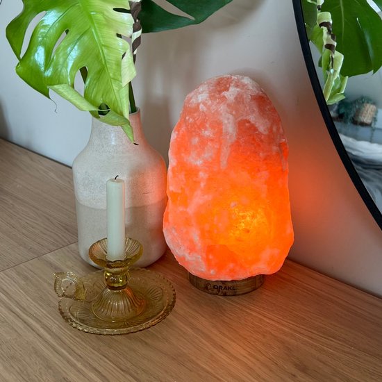 Een oranje gloeiende Himalaya zoutlamp op een houten plank, naast een vaas met een plant en een gele kandelaar.