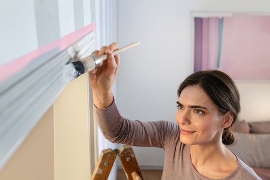 Vrouw met verfkwast schildert langs roze schilderstape op gestreepte muur met sierlijst.