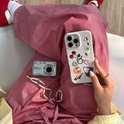 Vrouw in gestreepte broek houdt iPhone 15 "It Girl" hoesje vast, naast camera en parelkettingen.