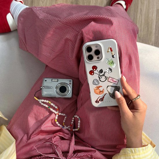 Vrouw in gestreepte broek houdt iPhone 15 "It Girl" hoesje vast, naast camera en parelkettingen.