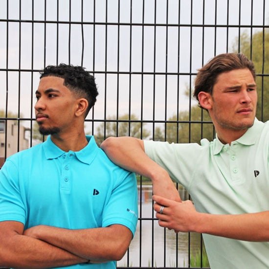 Twee mannen buiten bij een hek, één in turquoise polo, de ander in lichtgroene polo.