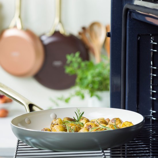 Lichtgekleurde aardappelen met rozemarijn in een bruine koekenpan met gouden handvat, op een rooster boven een oven.