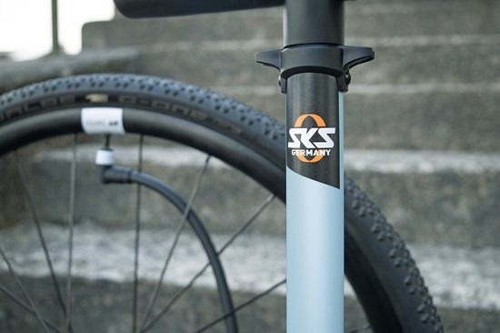 Close-up van de lichtblauwe cilinder van een fietspomp met het "SKS GERMANY" logo, naast een fietswiel.