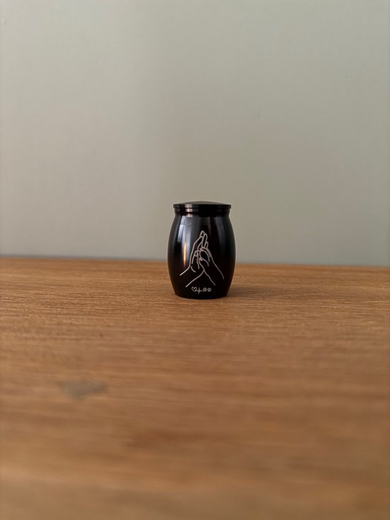 Zwarte mini urn met pootafdruk opdruk, staand op een houten tafel.