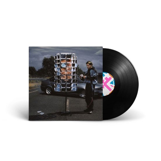 Een vinyl singlehoes met een persoon die een brief leest naast een glimmende, vierkante installatie op een landweg.