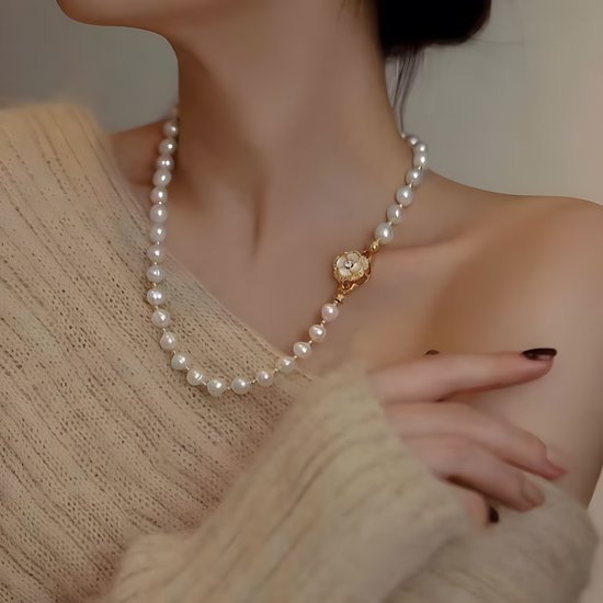 Crèmekleurige parelketting met een gouden bloemvormige sluiting, gedragen door een vrouw met een beige trui.