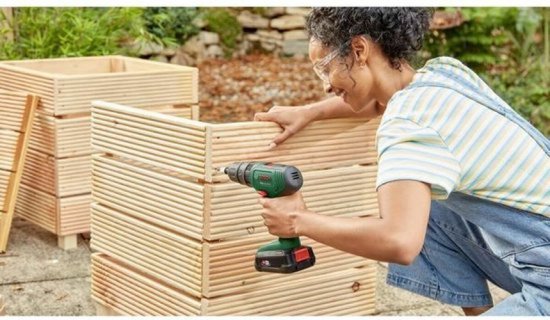 Vrouw met veiligheidsbril boort met een groene Bosch accuboorschroevendraaier in een houten plantenbak.