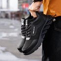 Paar zwarte werksneakers met mesh en stevige zolen, vastgehouden door een persoon.