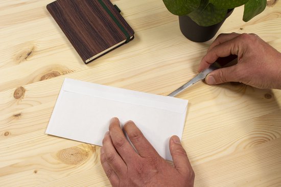 Handen openen een envelop met een briefopener op een houten bureau. Een notitieboekje en plant staan ernaast.