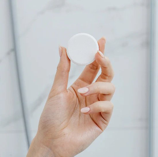 Hand met een naturel gelakte nagel houdt een witte, ronde douchetablet vast.