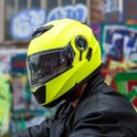 Man met een gele systeemhelm met zonnevizier, draagt een zwarte leren jas, graffiti op achtergrond.