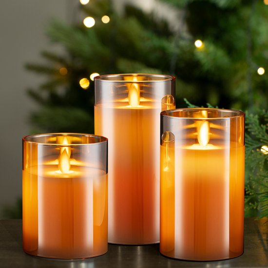 Drie amberkleurige LED-kaarsen in glazen potten met realistische, bewegende vlammen, geplaatst voor een kerstboom.