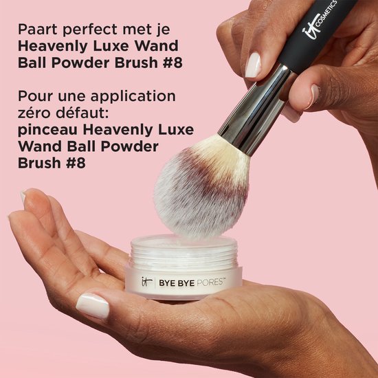 Een IT Cosmetics poederkwast met witte en bruine haren wordt boven een open potje Bye Bye Pores poeder gehouden.