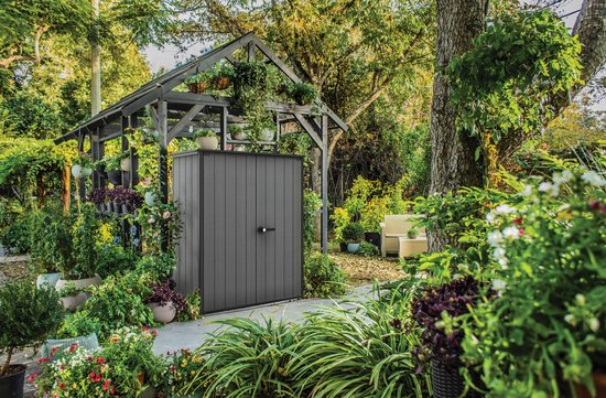 Abri de jardin gris dans un jardin luxuriant, à côté d'une pergola en bois avec des plantes et une chaise.