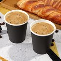 Twee zwarte papieren bekers met koffie en schuim, naast een croissant.