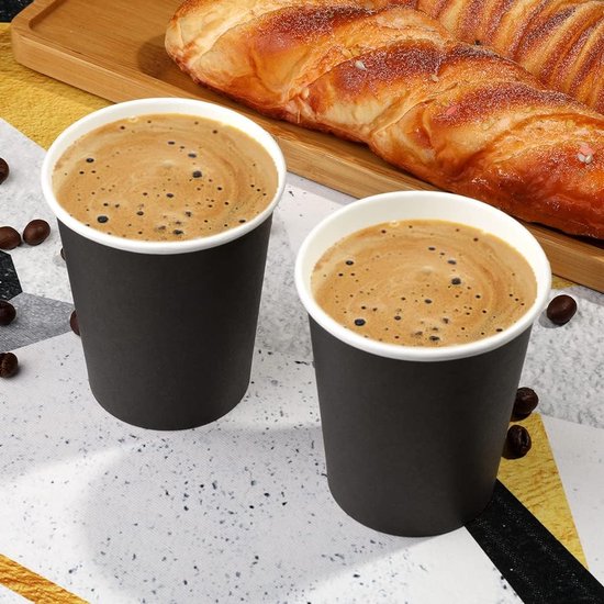 Twee zwarte papieren bekers met koffie en schuim, naast een croissant.