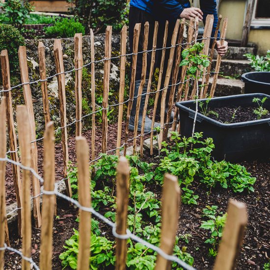 Man plaatst een houten schutting in de tuin. Palen van naturel hout verbonden met metaaldraad.