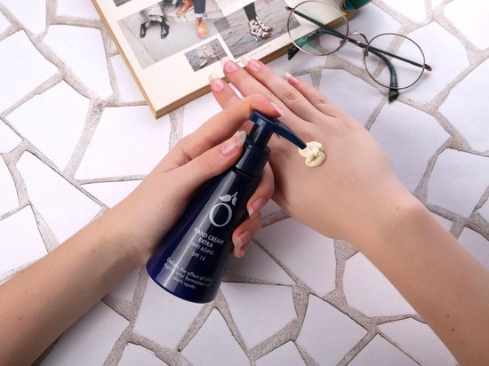 Handen met lange nagels, aangebrachte handcrème op de huid vanuit een blauwe pompflacon.