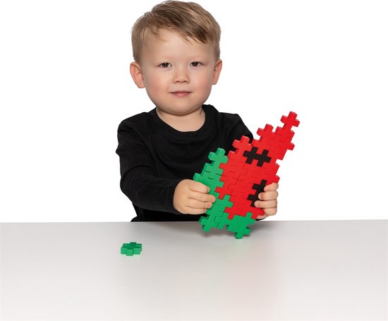 Een jongen met lang zwart shirt houdt een watermeloenvormige constructie vast, gemaakt van rode en groene Plus-Plus stukken.