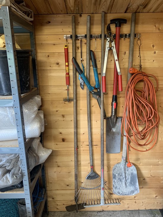 Gereedschap, tuingereedschap en slangen op een zwarte bezemhouder in een schuur.