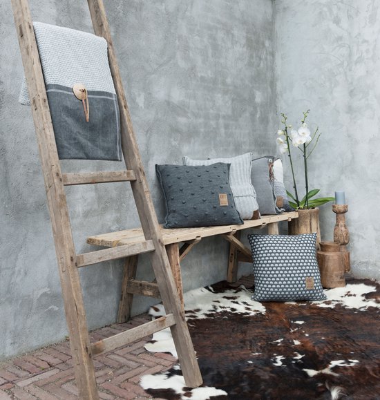 Grijze sierkussens op een houten bankje, met een gebreide lichtblauwe plaid op een ladder.