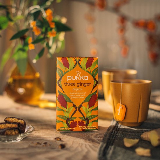 Pukka three ginger biologische thee verpakking op een houten tafel, met een mok en koekjes.