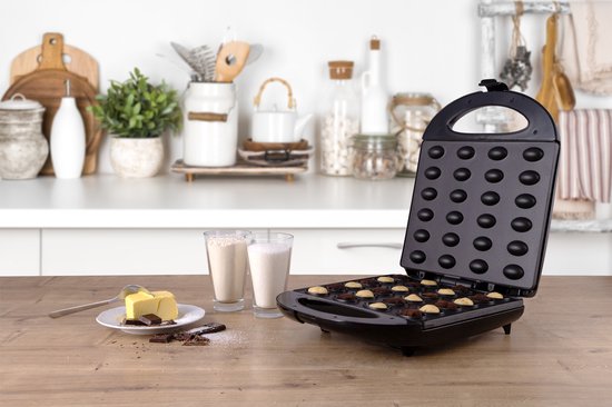 Zwarte koekjesmaker geopend met 24 ronde bakvormpjes, met ingrediënten op een houten tafel.
