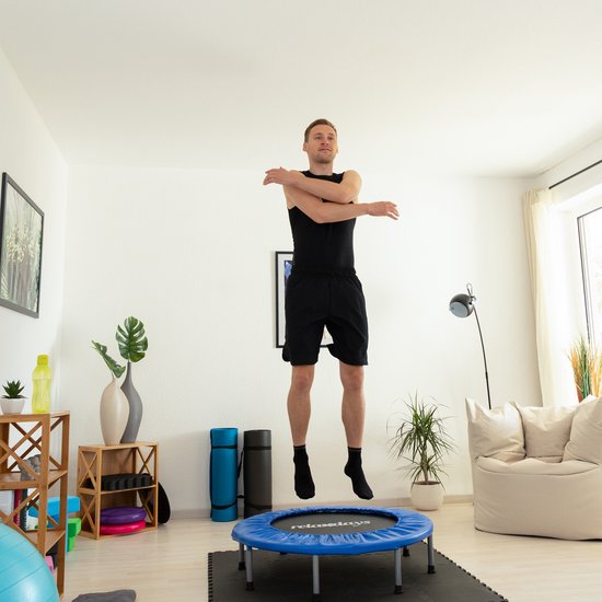 Een man in een zwart sportshirt en korte broek springt op een blauwe trampoline in een woonkamer.