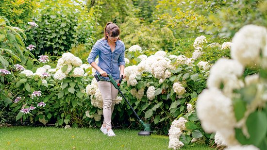Vrouw in spijkerblouse trimt gras in een tuin met witte hortensia's, gebruikmakend van de Bosch grastrimmer.