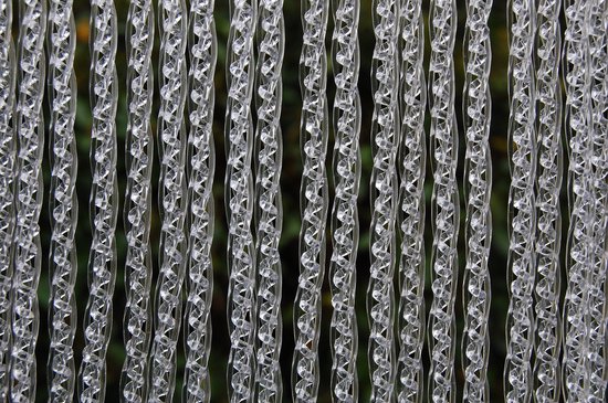 Een transparant deurgordijn met verticale, gedraaide plastic kettingen. Op de achtergrond vaag groen van de natuur.